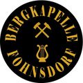 Ein rundes Logo mit 'Bergkapelle' und 'Johannsdor' in Gold auf schwarzem Hintergrund, mit einem Leier- und Spitzhackensymbol.