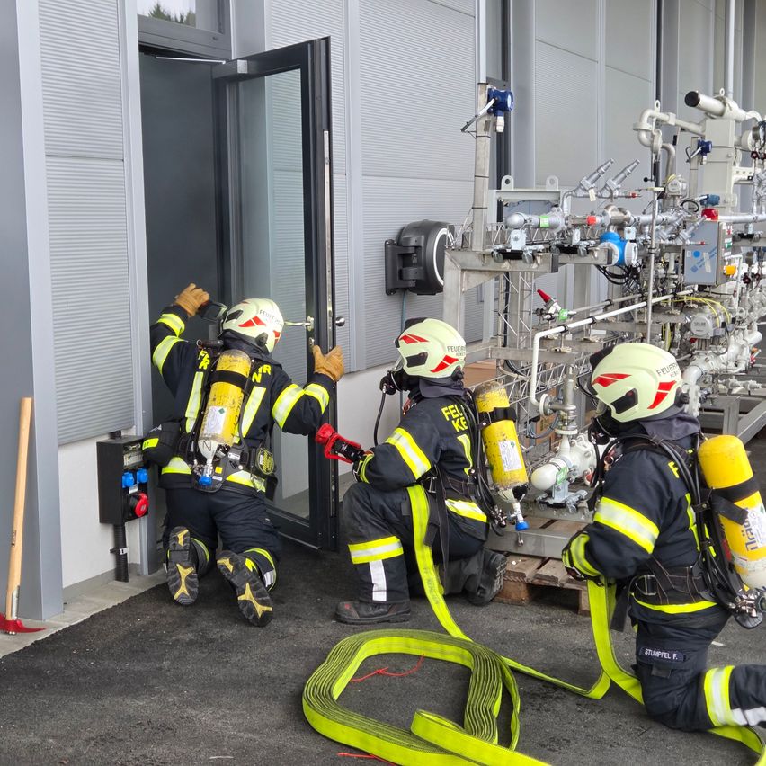 Three firefighters in full gear prepare to enter a building, with one opening the door and two others ready with a hose and equipment.