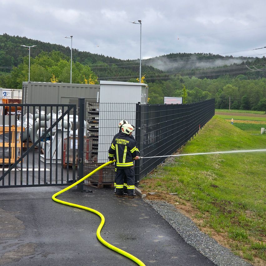A firefighter is spraying water from a hose at a fence, near stacked gas cylinders and a gate. Trees and mountains are in the background.
