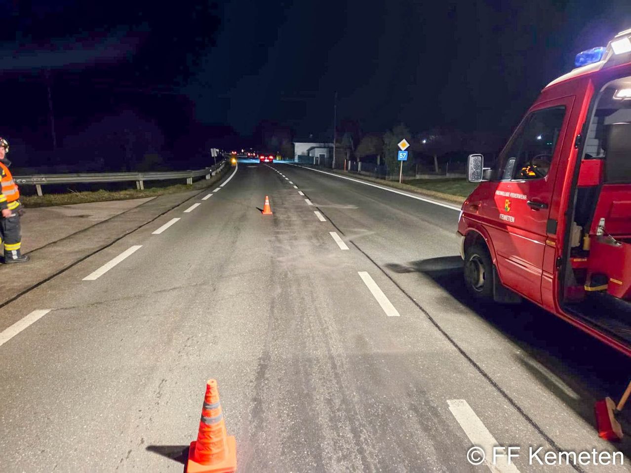A nighttime road scene with a red vehicle on the right and traffic cones on the left. The road has white markings and a sign with the number 57 on the right.