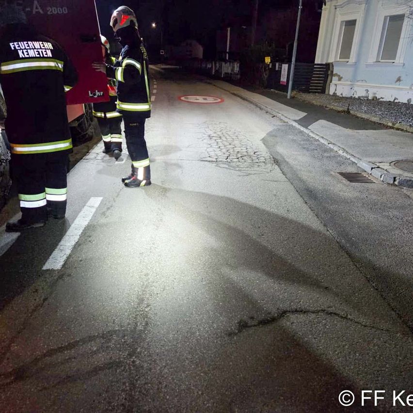Firefighters are on the road at night, with one holding a red box. The road has a curb and a small circle on the road.