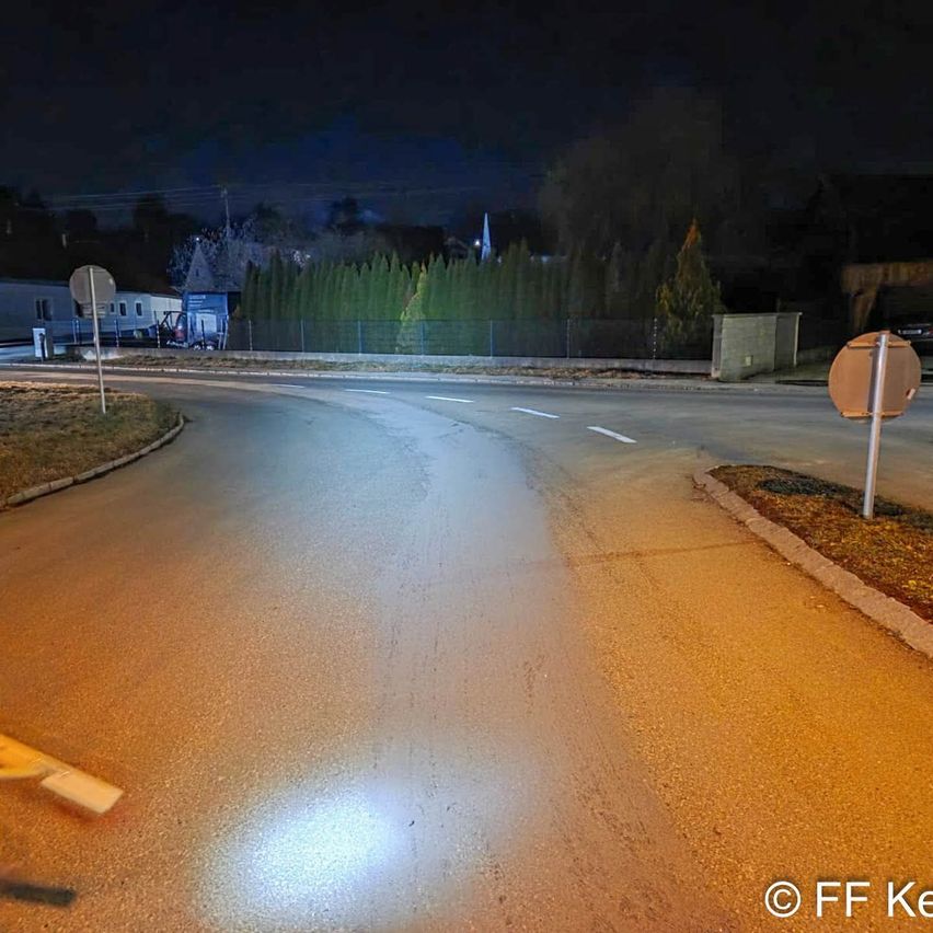An empty street at night with a car light reflecting on the road. There are street signs and a pole on the side.