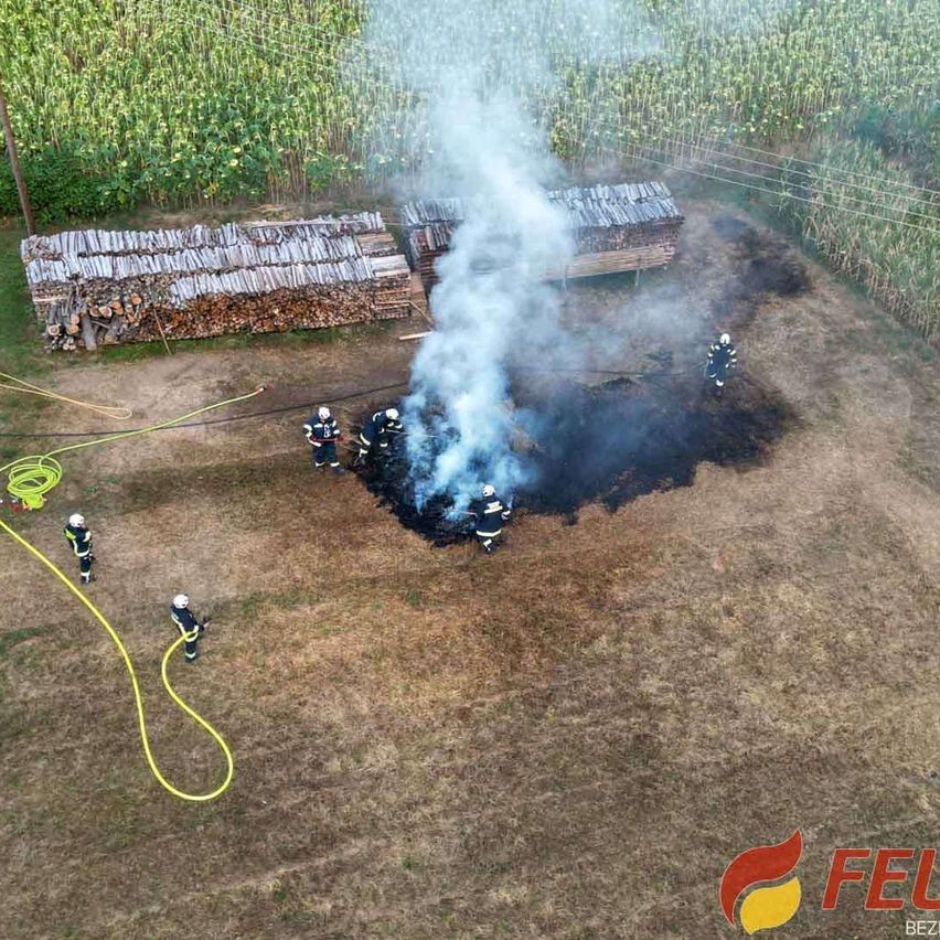 Feuerwehrleute löschen Brand bei Holzstapeln in Feldnähe, Rauch steigt auf.