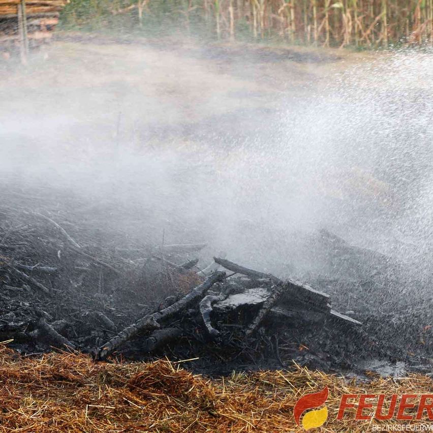 Feuerwehr löscht brennenden Holzhaufen mit Wasserstrahl in ländlicher Umgebung.