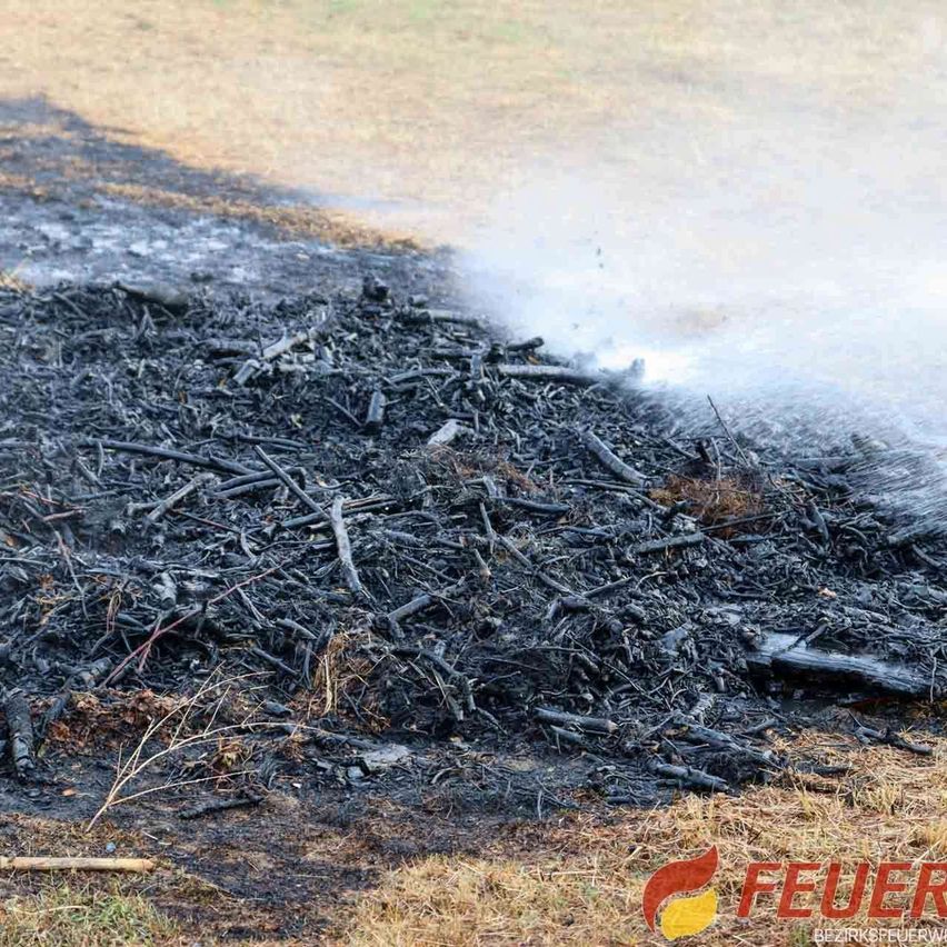 Ein Haufen verkohlter Holz- und Zweige wird mit Wasser übergossen, wahrscheinlich um ein verbleibendes Feuer zu löschen. Der Boden ist mit Asche bedeckt.