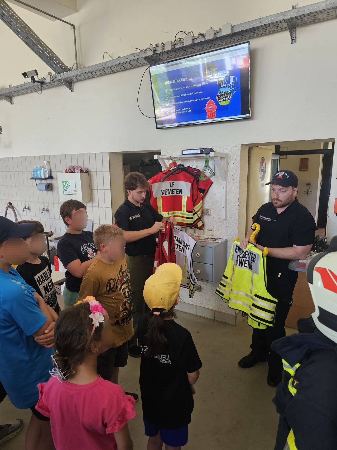 Zwei Feuerwehrleute zeigen einer Gruppe von Kindern in einer Feuerwache Rettungswesten.