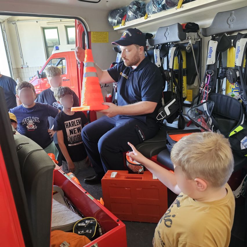 Ein Feuerwehrmann zeigt Kindern im Inneren eines Feuerwehrautos. Ein Kind zeigt, während andere einen Kegel beobachten.