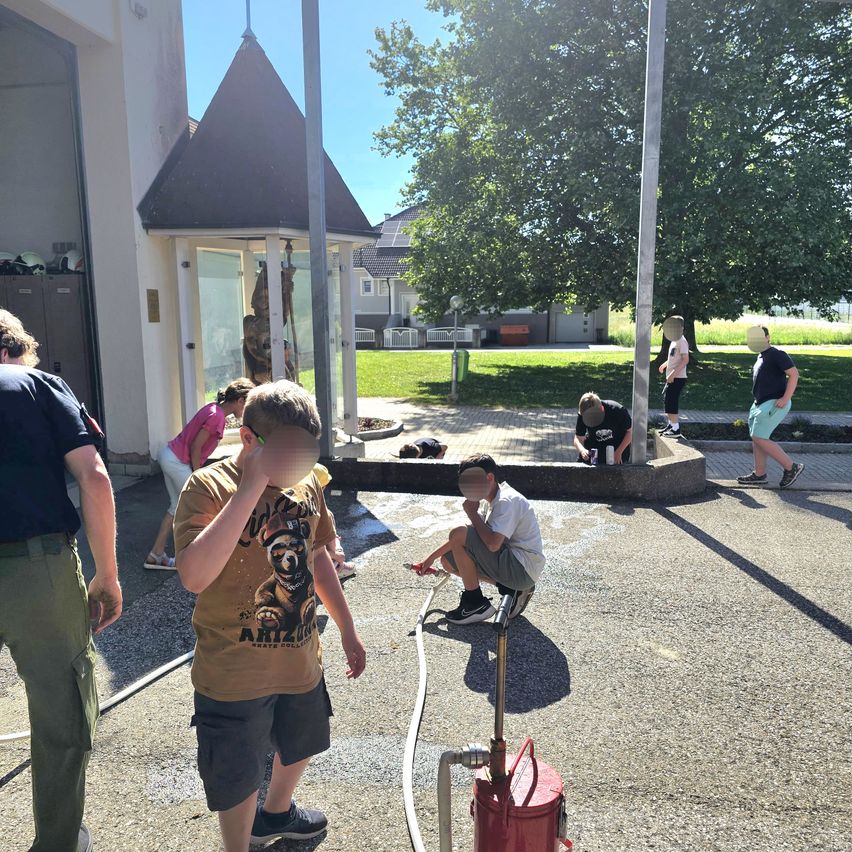 Eine Gruppe junger Menschen ist draußen, einige putzen mit Schläuchen. Ein Junge kniet auf dem Boden, möglicherweise zur Reinigung einer Statue. Ein Feuerlöscher ist in der Nähe.