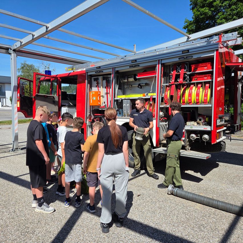 Eine Gruppe von Kindern beobachtet zwei Feuerwehrleute in einem roten Feuerwehrauto. Sie stehen unter einer Metallstruktur, während die Feuerwehrleute etwas erklären.
