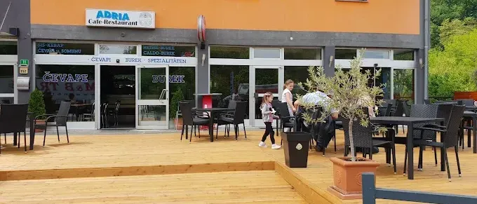 Der Außenbereich des Adria Cafe-Restaurants verfügt über Holztische und -stühle, einen Topf mit Olivenbaum und ein Kind, das vorbeigeht. Das Gebäude hat Glastüren und -fenster und ein Schild über dem Eingang.