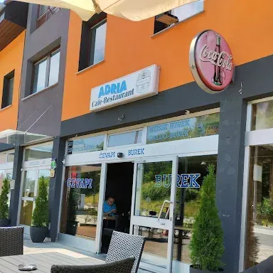 Ein Restauranteingang mit einem Coca-Cola-Schild, einer Person, die an einem Tisch drinnen sitzt, und Außentischen mit Stühlen davor.