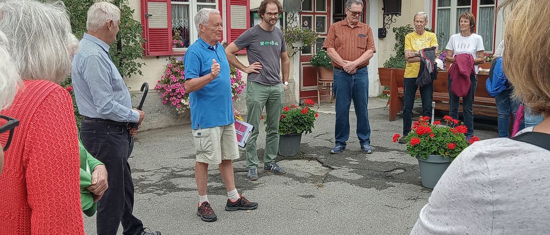 Eine Gruppe von Menschen, einige in Freizeitkleidung, versammelt sich vor einem Gebäude mit roten Fensterläden und Blumen, möglicherweise lauschen sie einem Sprecher.