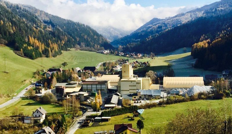 Ein Luftbild eines Dorfes zwischen Bergen, mit einem großen Industriegebäude und Häusern, eingebettet in einem üppigen grünen Tal unter einem teilweise bewölkten Himmel.