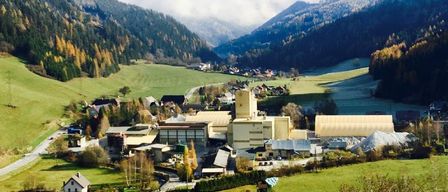 Ein Luftbild eines Dorfes zwischen Bergen, mit einem großen Industriegebäude und Häusern, eingebettet in einem üppigen grünen Tal unter einem teilweise bewölkten Himmel.