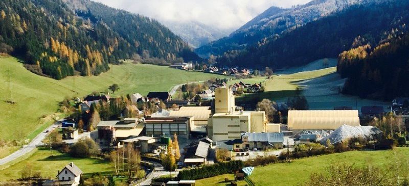 An aerial view of a village nestled between mountains, with a large industrial building and houses, set in a lush green valley under a partly cloudy sky.