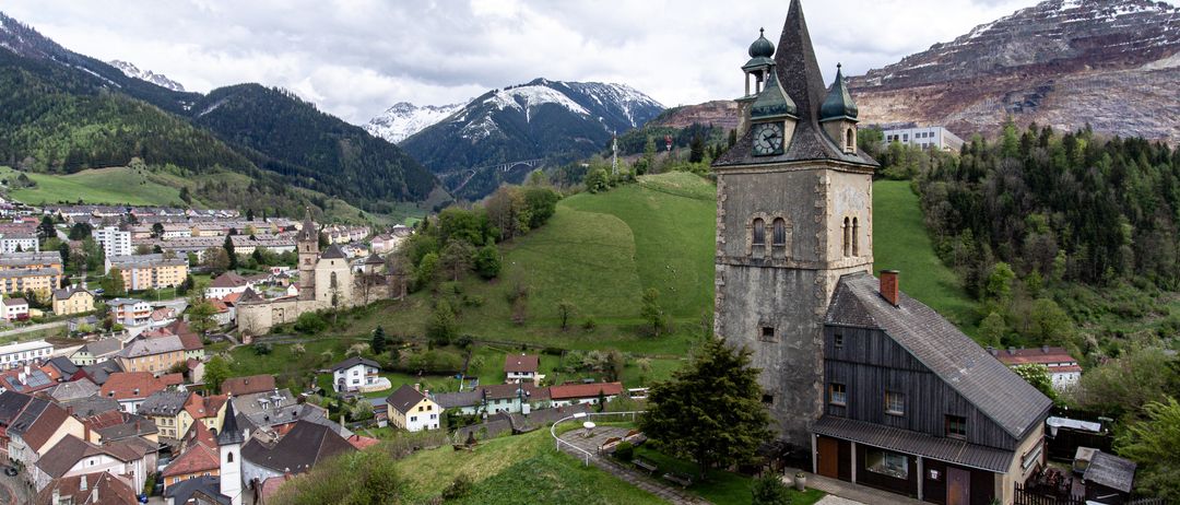 Eine Luftaufnahme einer Stadt mit einem Uhrturm, umgeben von grünen Hügeln und schneebedeckten Bergen in der Ferne.