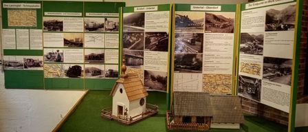 Eine Miniatur-Zugausstellung zeigt einen Modellbahnhof, umgeben von verschiedenen Fotos, Karten und Texttafeln. Ein kleines Holzhaus und eine Miniaturkirche sind ebenfalls vorhanden.