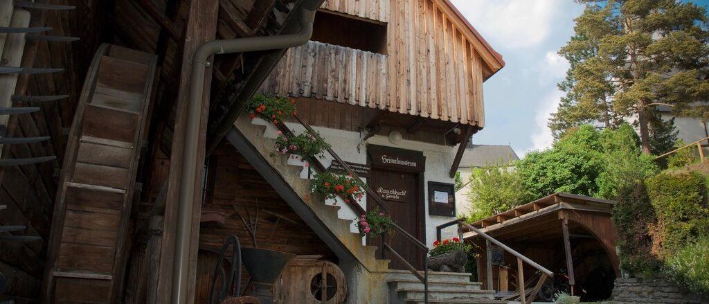 Ein Holzhaus mit Stufen zum Eingang, geschmückt mit Blumen. Eine rustikale Holzkonstruktion und Bäume im Hintergrund.