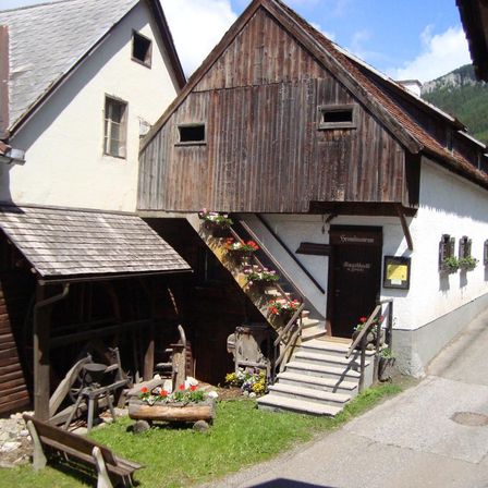 Ein rustikales Haus mit holzverkleideter Außenseite und Stufen, die zum Eingang führen. Es hat mehrere Fenster und eine Tür mit Namensschild. Blumentöpfe und Blumen schmücken die Vorderseite.