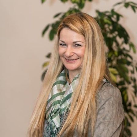 Eine lächelnde Frau mit langen blonden Haaren steht neben einer Pflanze und trägt einen grauen Mantel und einen grünen Schal.