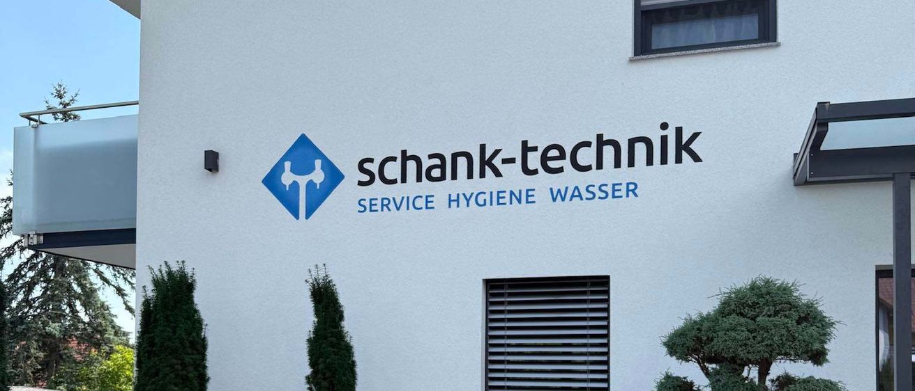 Ein weißes Gebäude zeigt das Logo und den Text 'Schank-Technik Service Hygiene Wasser'. Ein Fenster mit Rollladen befindet sich über dem Logo. Der Eingang hat einen überdachten Weg. Vor dem Gebäude stehen Topfpflanzen und Sträucher.
