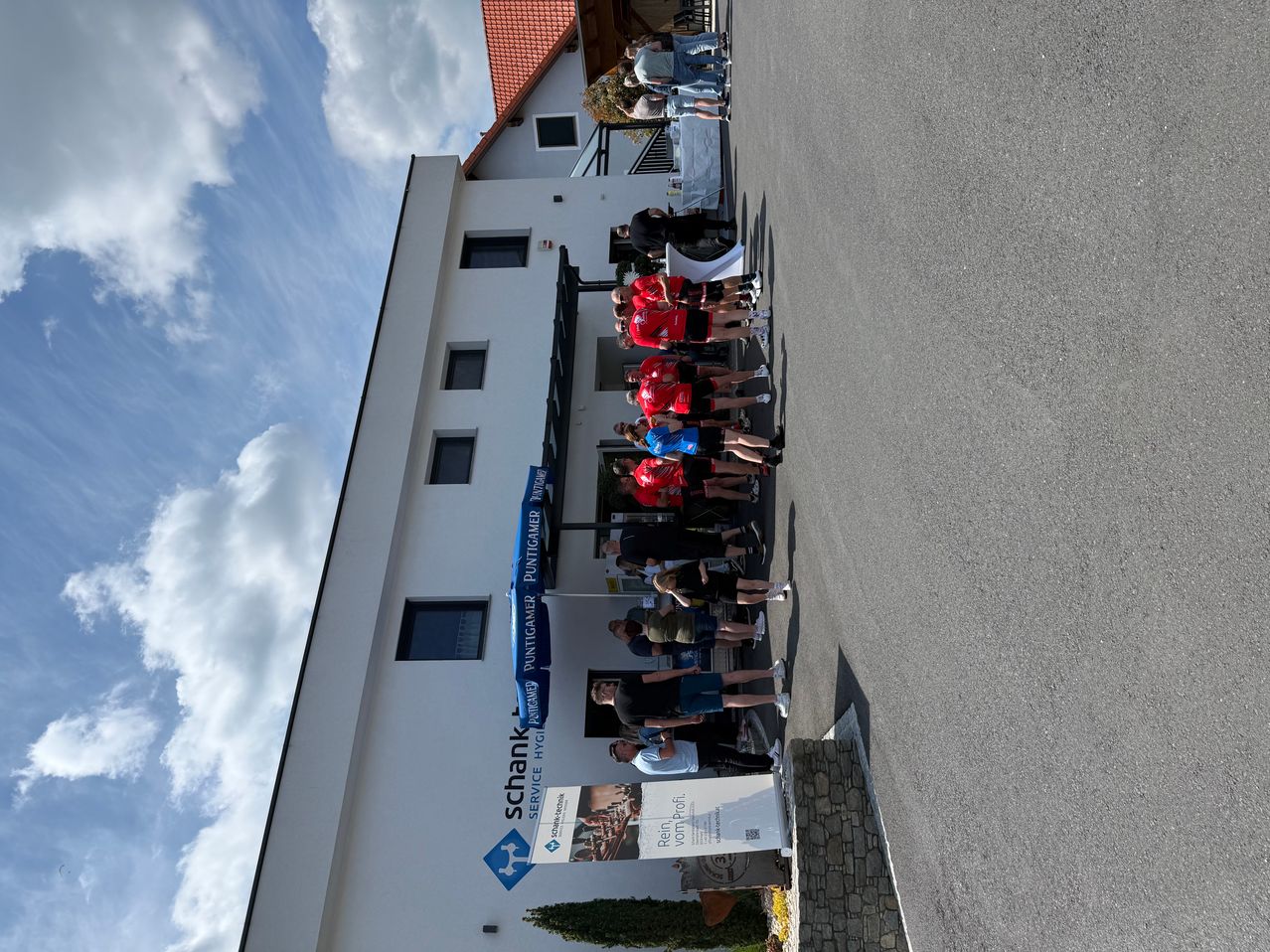 Eine Gruppe von Menschen in roten Shirts steht in einer Reihe vor einem weißen Gebäude. Das Gebäude hat ein Schild mit der Aufschrift 'Schank Service' und eine blaue Fahne.