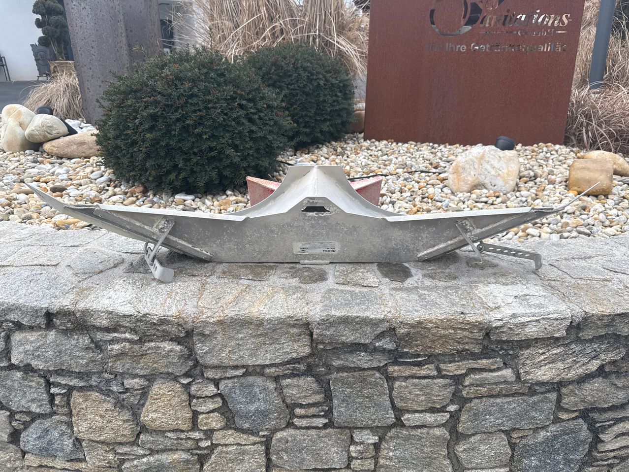 Eine Metallskulptur, die einem Schiff ähnelt, ist auf einer Steinmauer mit Steinen und Pflanzen im Hintergrund platziert.