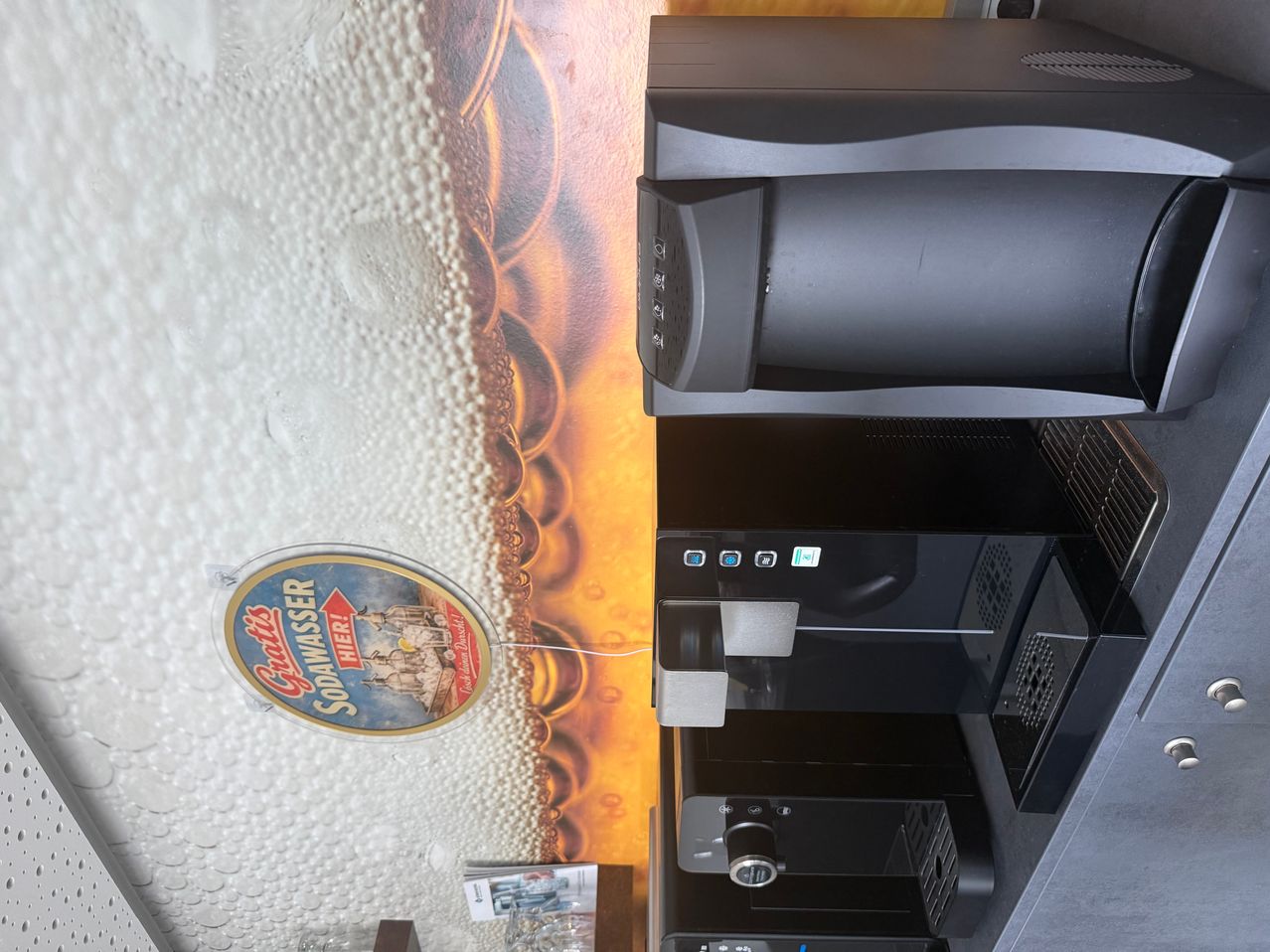 A black coffee machine is shown from the side, with a decorative beer glass design on the left. The machine has buttons and a screen, and it appears to be in a bar or restaurant.