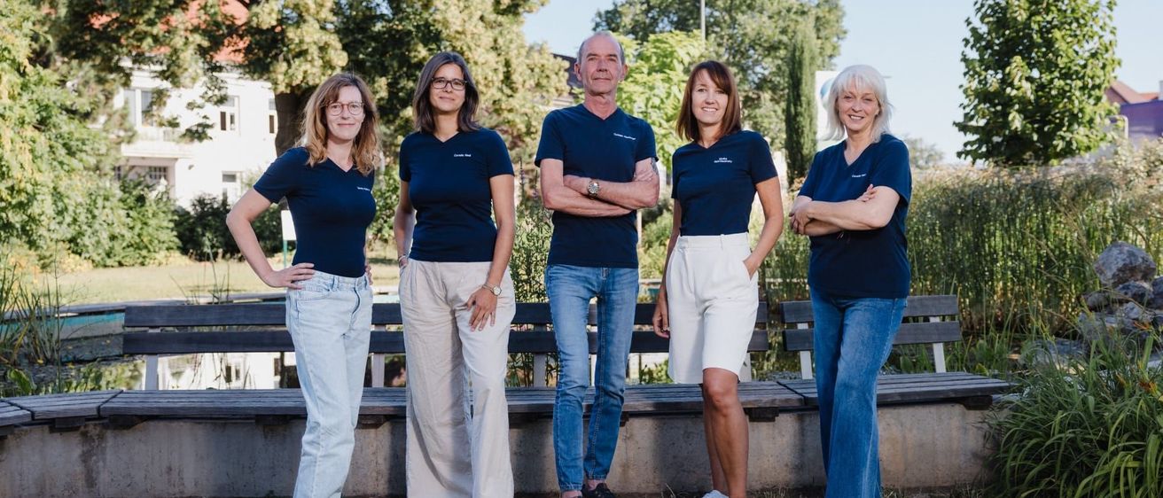 Fünf Menschen stehen in einem Park zusammen und tragen identische blaue T-Shirts. Sie haben die Arme verschränkt und lächeln für die Kamera. Hinter ihnen sind Bänke und Bäume.