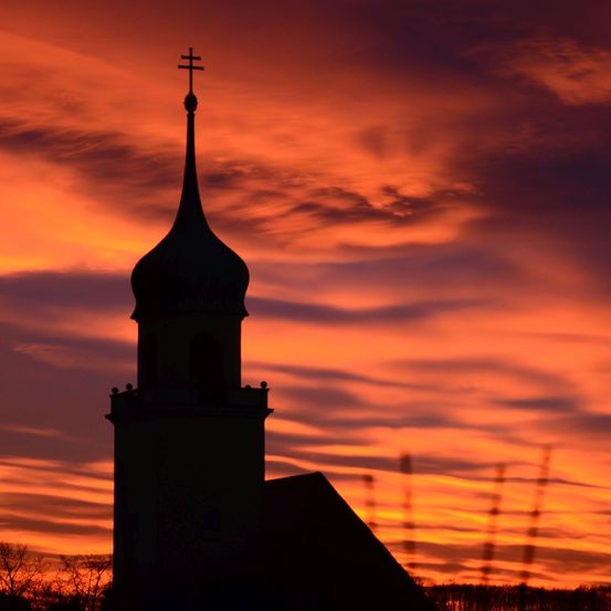 Die Silhouette eines Kirchturms mit einem Kreuz auf der Spitze, vor dem Hintergrund eines orange-pinken Sonnenuntergangs.