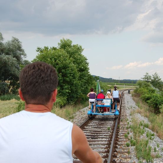 Ein Mann beobachtet eine Gruppe von Menschen, die auf einem pedalbetriebenen Wagen auf einem Bahngleis fahren, mit Grünpflanzen und einem bewölkten Himmel im Hintergrund.