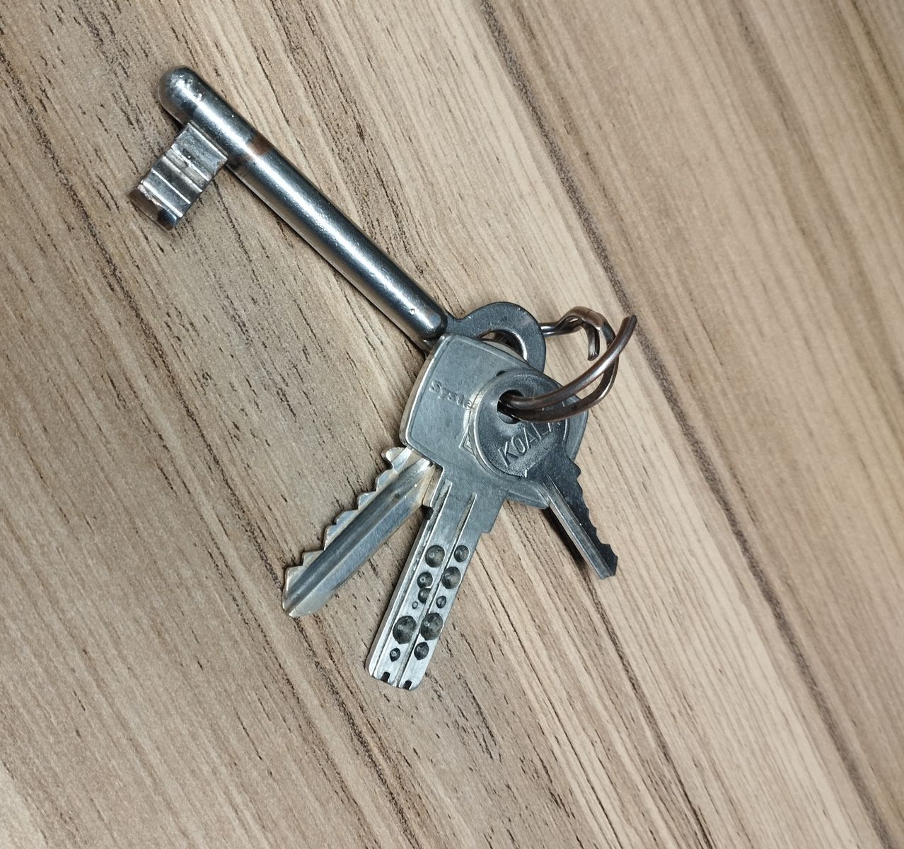 Ein Satz silberner Schlüssel auf einer Holzoberfläche. Die Schlüssel haben Metallköpfe und Kunststoffkörper, wobei ein Schlüssel einen kleinen Ring aufweist.