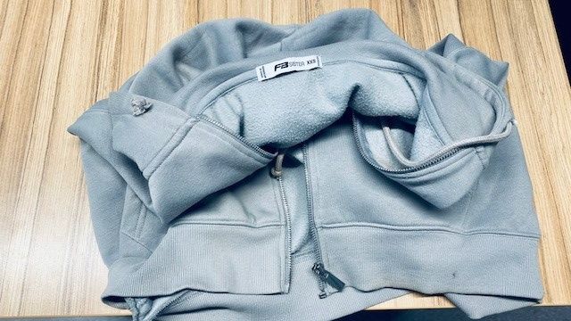 Eine hellblaue Kapuzenjacke mit Reißverschluss liegt flach auf einer Holzoberfläche. Die Jacke hat eine Kapuze und Taschen, und der Reißverschluss ist teilweise geöffnet.