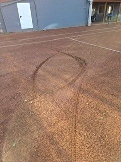 An empty tennis court with tire tracks on the surface and a building with a white door in the background.