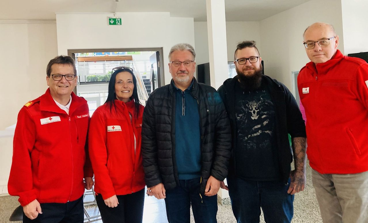 Vier Personen, zwei Männer und zwei Frauen, stehen in einem Flur. Die Frau auf der linken Seite trägt eine rote Jacke mit einem weißen Logo. Der Mann in der Mitte trägt eine Brille und eine dunkle Jacke. Der Mann auf der rechten Seite hat einen Bart und trägt eine schwarze Kapuzenjacke. Hinter ihnen befindet sich eine offene Tür.