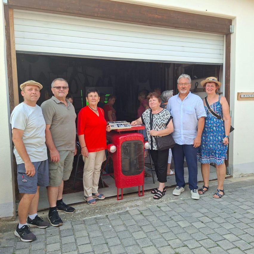 A group of people stands in front of a garage, with a red fan and a table in the middle. They are smiling and appear to be posing for a photo.