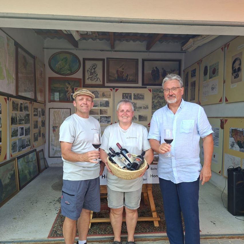 Three men stand in a room with a basket of wine bottles. They are smiling and holding wine glasses. The room has many framed pictures on the walls.