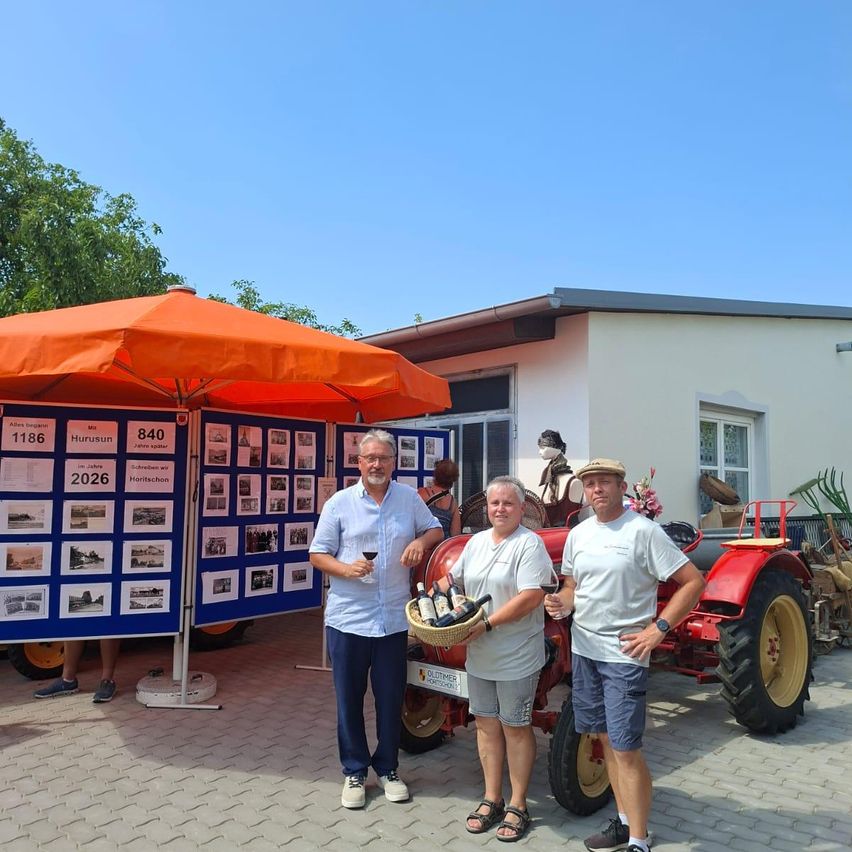 Three people stand in front of a red tractor, one holding a basket of wine bottles. They are smiling and appear to be posing for a photo. Behind them is a blue banner with pictures and text, and an orange umbrella.