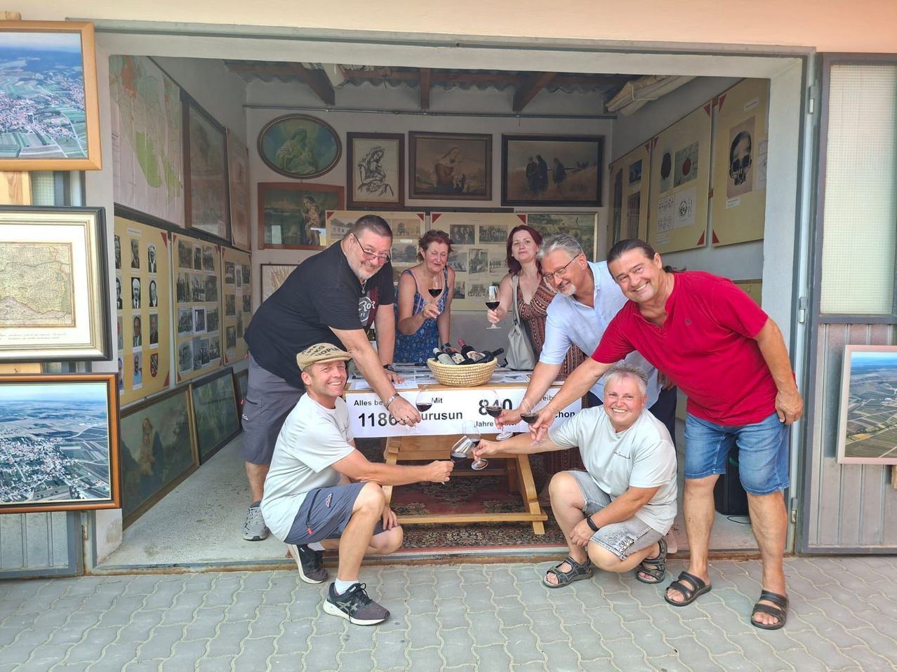 A group of people are gathered in a room, smiling and holding wine glasses. They are standing around a table with bottles and a basket. The room has a wall filled with framed pictures.