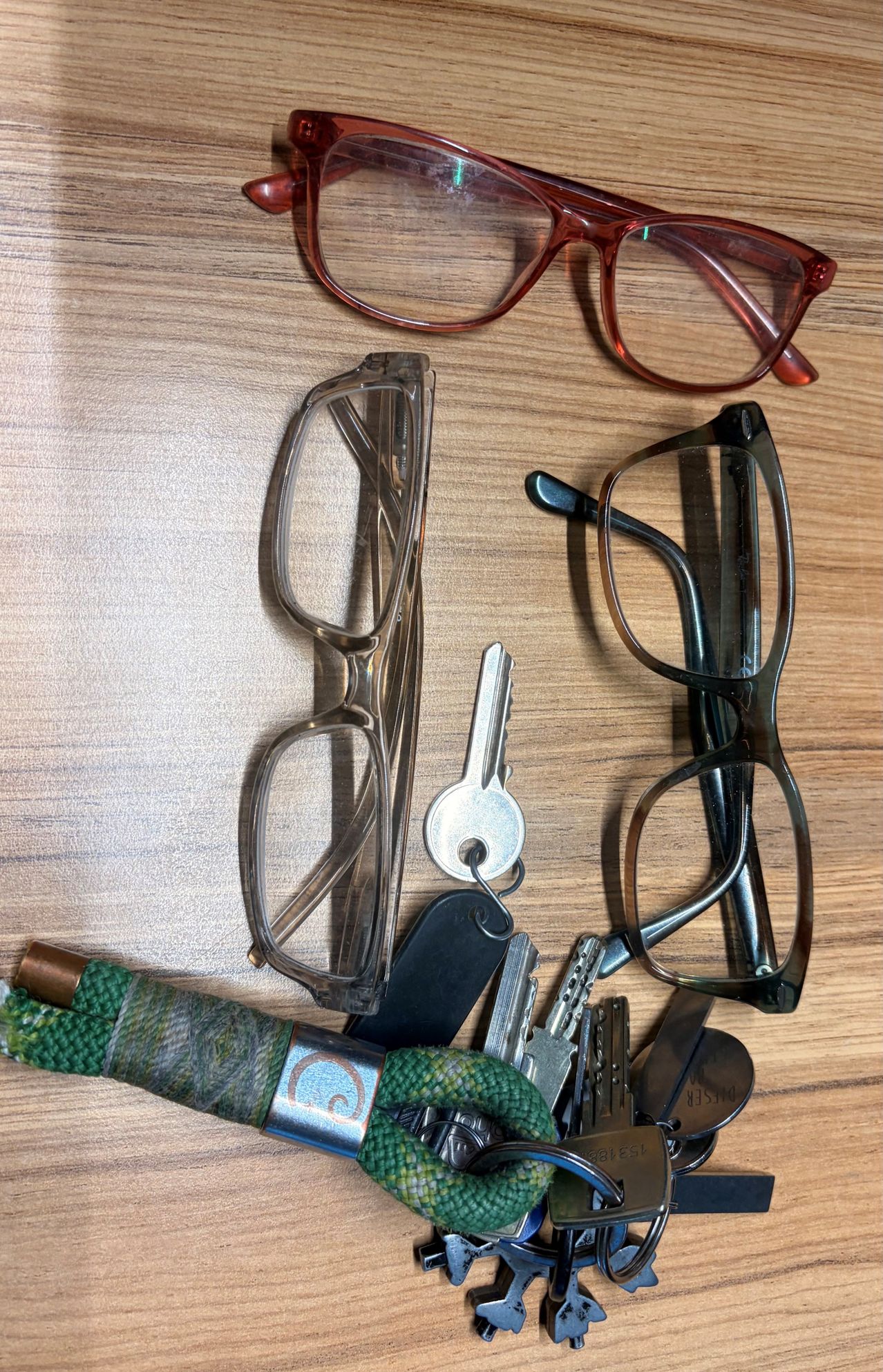 A wooden surface holds several eyeglasses, a keychain, and keys. The eyeglasses are clear with metal frames. The keychain is black, and the keys are silver.