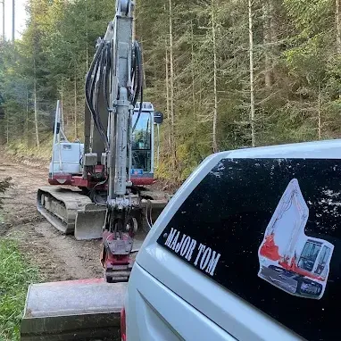 Ein großer Bagger ist auf einer unbefestigten Straße geparkt. Ein weißer Wagen mit einem Aufkleber, der 'MAJOR TOM' lautet, steht davor.