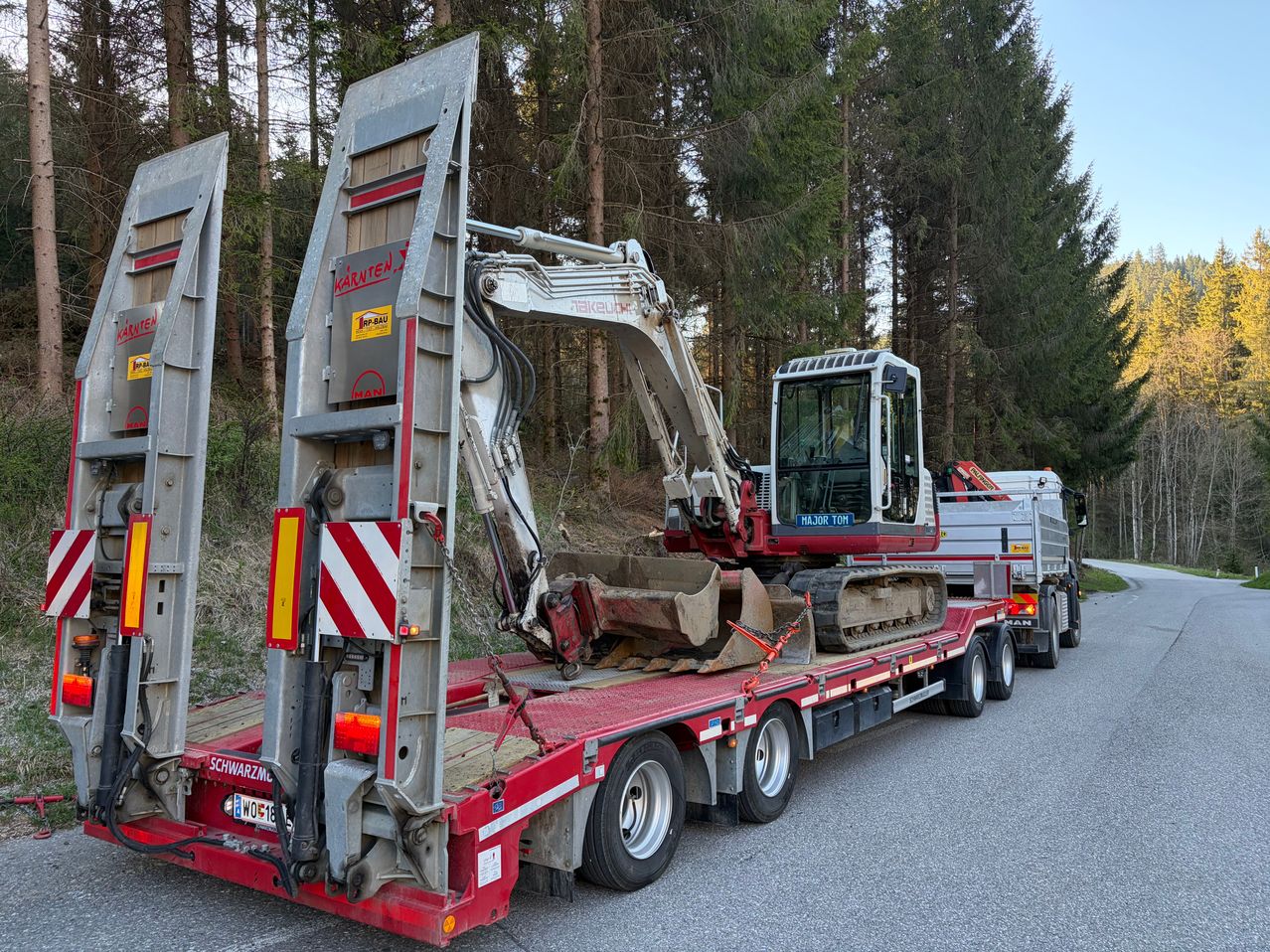 Ein roter Anhängertransporter transportiert einen Baggerlader auf einer Straße, umgeben von Bäumen. Der Lader ist mit Ketten gesichert und hat einen gelben Aufkleber an der Seite.