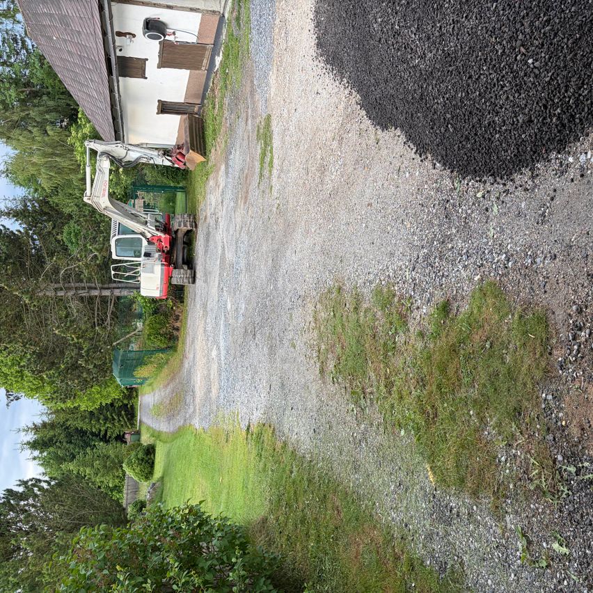 Ein Baustellenbereich mit Kies und einer rot-weißen Raupe auf der linken Seite. Ein Haus befindet sich im Hintergrund.