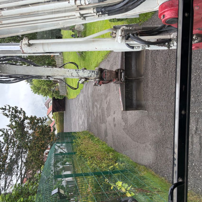 Blick von innen aus einem Fahrzeug auf eine große Maschine mit einem roten Teil auf einem Kiesweg. Bäume und ein grünes Gitter sind zu sehen.
