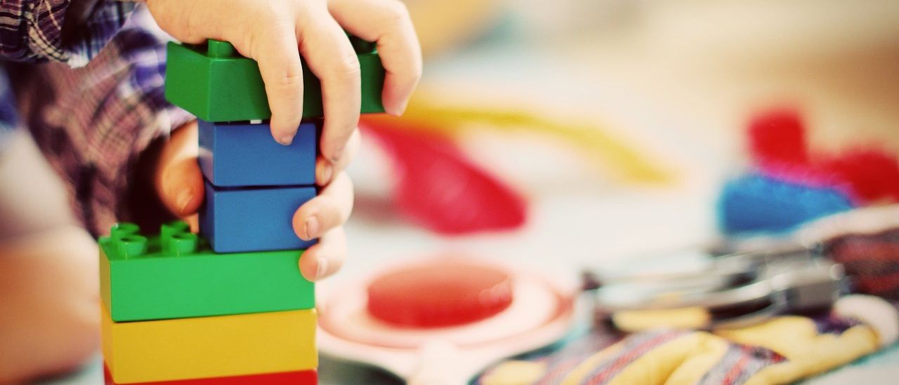 Ein Kind baut bunte Bausteine auf einem Tisch, mit einem weißen Teller und Handschuhen in der Nähe.