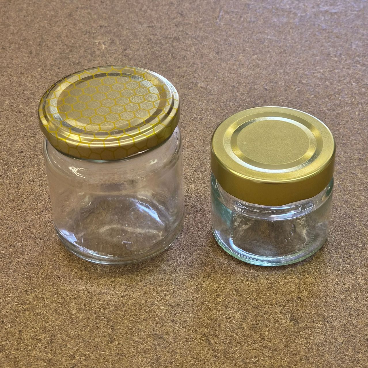Two clear glass jars with gold lids, one with a hexagonal pattern on the lid, sit on a brown surface. Both jars are empty.