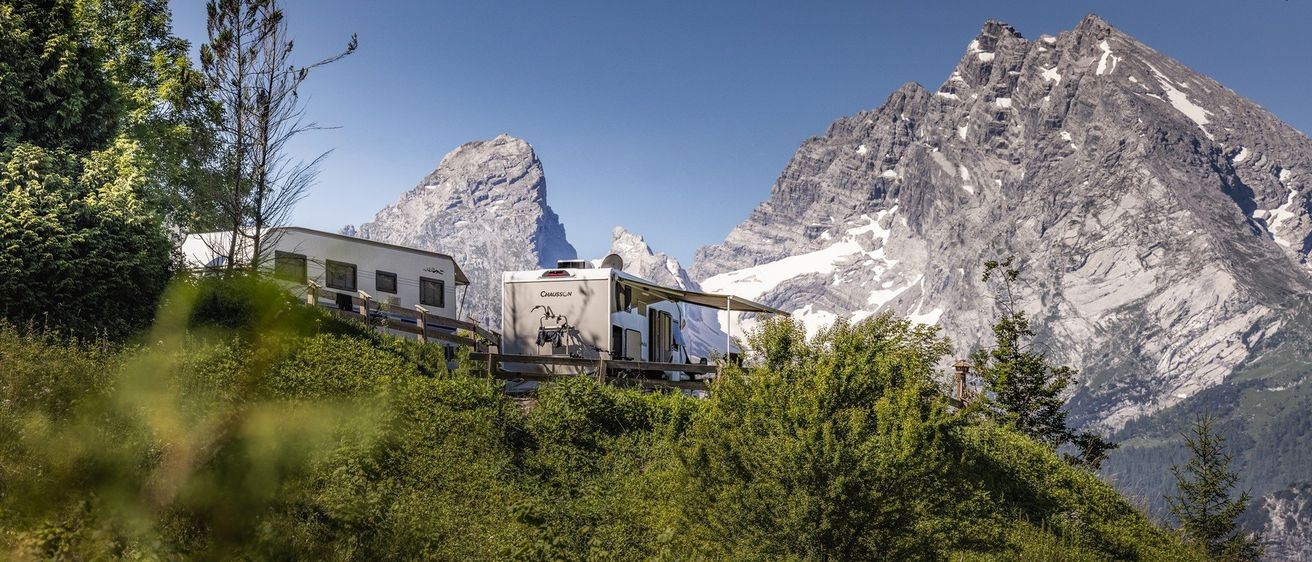 Zwei Wohnwagen, die zwischen Bäumen geparkt sind, mit Bergen im Hintergrund. Der Himmel ist blau.
