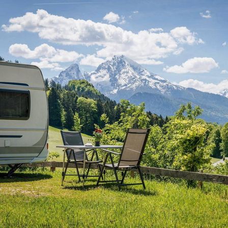 Ein weißer Wohnwagen auf einem Rasenfeld mit Tisch und Stühlen. Berge im Hintergrund mit Schnee oben.