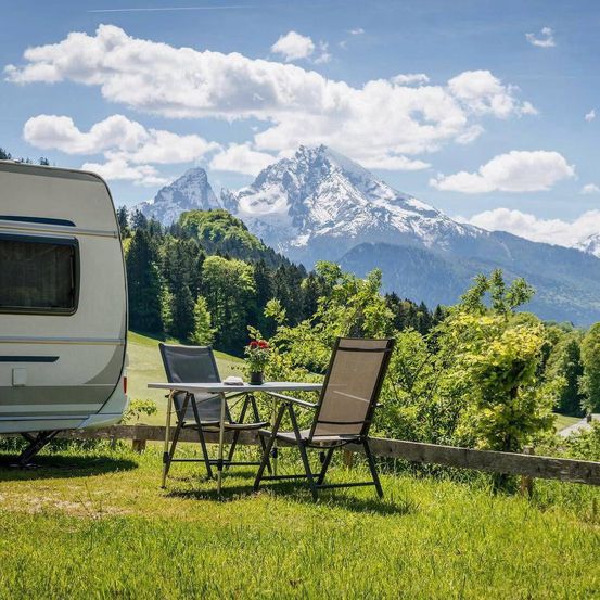 Ein weißer Wohnwagen auf einem Rasenfeld mit Tisch und Stühlen. Berge im Hintergrund mit Schnee oben.