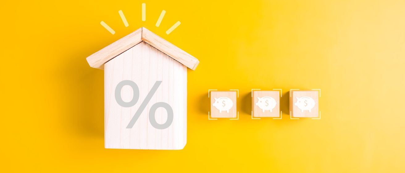 Ein hölzernes Hausmodell mit einem Prozentzeichen auf gelbem Hintergrund und drei Sparschweinmodellen mit Dollarzeichen auf der rechten Seite.